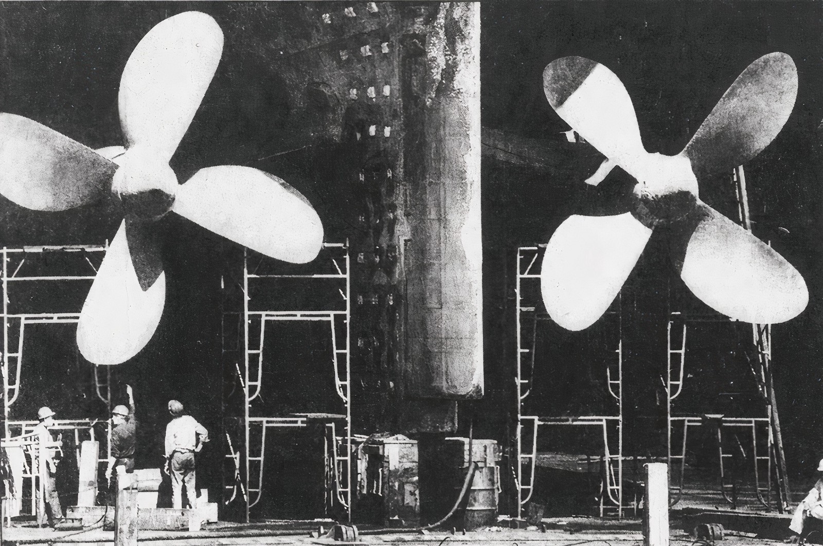 Black-and-white photo of two giant ship propellers towering over workers on scaffolding at the East Boston shipyard