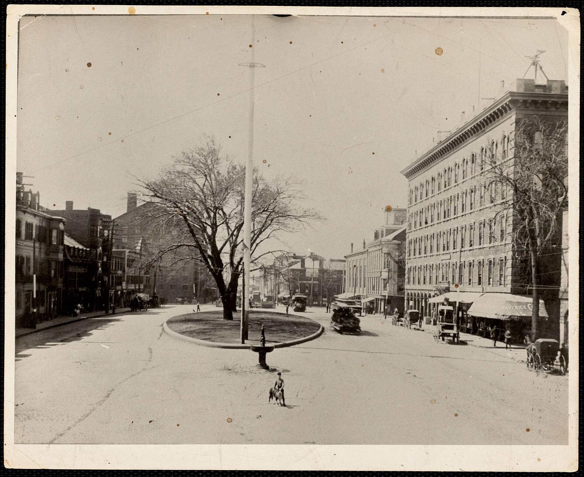 Historic photo of Maverick Square in East Boston with a small green, a flagpole and brick commercial buildings